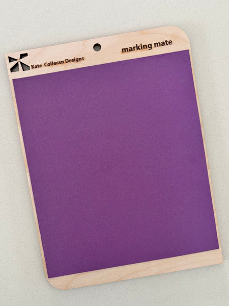 A purple marking mat with a light natural wood border and a hanging hole, branded 'Kate Coleran Designs' and labeled 'marking mate'.