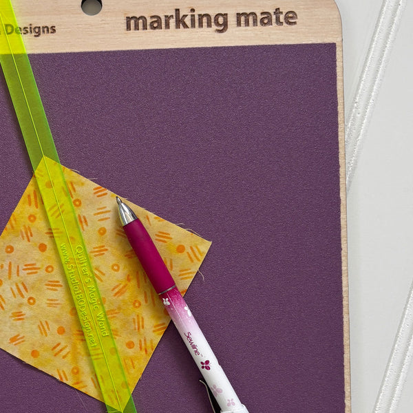 A close-up of a purple marking mat in its wooden hanger packaging, with a pink and white Sakura pen and a yellow patterned square card placed on top.
