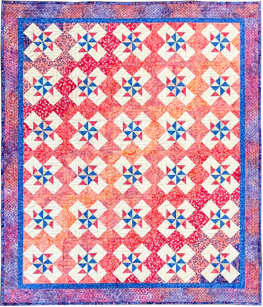A square quilted textile with a repeating geometric star and diamond block pattern in coral, pink, orange, blue, and cream, finished with a blue patterned border.